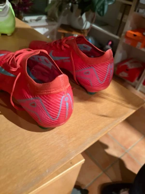 Röda Nike fotbollsskor med blå swoosh - Säljer ett par röda Nike fotbollsskor med stor blå swoosh på sidan. Skorna har snörning och är tillverkade i ett lätt syntetmaterial som ger bra känsla på planen. Perfekta för dig som vill sticka ut på fotbollsplanen. Använda 5 gånger Stl 44