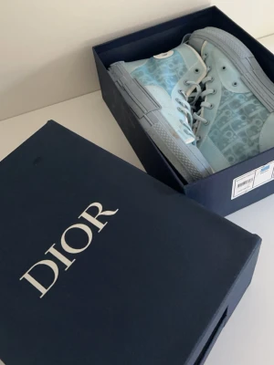 Blå Dior sneakers med monogram - Säljer ett par exklusiva blå Dior sneakers med det klassiska Dior-monogrammet på sidan. Skorna har snörning, rund tå och platt sula. Kommer i originalkartong med tillhörande certifikat och dustbag. Perfekta för dig som vill ha något unikt och trendigt.