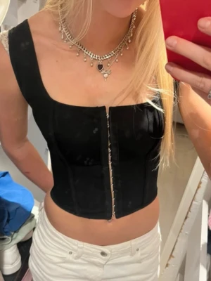 Svart korsett-topp med snörning - Snygg svart korsett-topp med breda axelband och croppad passform. Toppen har en dekorativ dragkedja framtill och snörning i ryggen för en justerbar passform. Perfekt att styla med både jeans och kjol för en trendig look.
