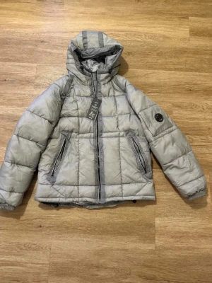 Grå pufferjacka från CP Company - Säljer en grå pufferjacka från CP Company med huva och två dragkedjeförsedda fickor. Jackan har en ikonisk linsdetalj på ärmen och quiltad design. Perfekt för kalla dagar och har en modern, sportig look.