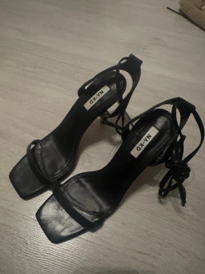 Svarta sandaletter med snörning från NA-KD - Säljer ett par stilrena svarta sandaletter från NA-KD med fyrkantig tå och tunna remmar som knyts runt ankeln. Skorna har klack och är tillverkade i ett slätt material som liknar skinn. Perfekta för dig som gillar minimalistisk och trendig stil. Använda 1 gång!
