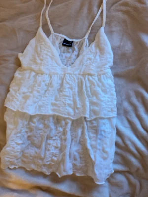 Vit volangig linnetopp från ginatrecot - Söt vit linnetopp från ginatrecot med tunna axelband och dubbla volanger. Toppen har v-ringning och är tillverkad i ett lätt, bomullstyg. Perfekt för varma dagar och enkel att styla med jeans eller kjol.