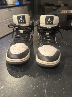 Nike Air Jordan 1 Mid svartvita sneakers - Snygga svartvita Nike Air Jordan 1 sneakers med klassisk design. Skorna har svart snörning, rund tå och vit sula. Tillverkade i skinn med ikoniska Jordan-loggan på plösen och sidan. Perfekta för dig som vill ha en stilren och tidlös look.