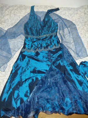 Mörkblå balklänning - Säljer en blå afton/balklänning med omlottkjol, v-ringad överdel och ett axelband. Finns två mindre band som man kan knyta till en blomma, samt en scarf att ha runt armarma. Storlek L, då midjan är ~84 cm (står M dock). Färgen är lite missvisande på första bilden, 2a visar lite bättre. Köptes begagnat och var för stor för mig. Bandet längst uppe behöver knappar, annars är den i bra skick. 