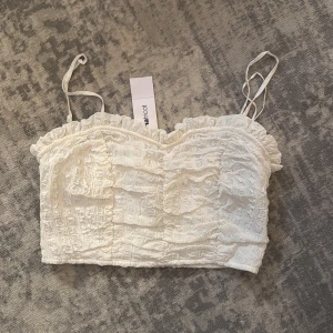 Vit croppad linnetopp med volang - Supersöt vit croppad linnetopp från Gina tricot med tunna axelband och volangkant upptill. Toppen har ett strukturerat tyg med rutigt mönster och smock baktill för extra komfort. Perfekt att styla med höga jeans eller kjol. Helt ny med prislapp kvar!