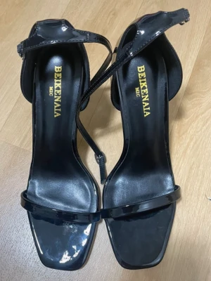 Svarta lackpumps från Bekenana - Stilrena svarta pumps från Bekenana i glansigt lackmaterial. Skorna har fyrkantig tå, smal rem över tårna och justerbar ankelrem. Perfekta för dig som gillar minimalistisk och trendig design med hög klack.