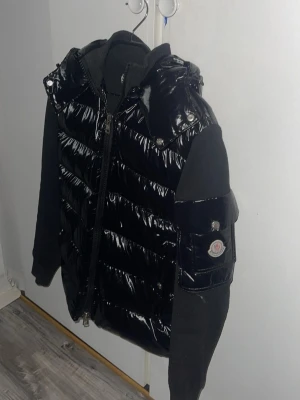 Svart glansig dunväst med huva - Snygg svart dunväst med glansig finish och huva. Västen har dragkedja framtill, två fickor med tryckknappar och breda quiltade paneler. Perfekt för lager på lager och streetwear-vibe.