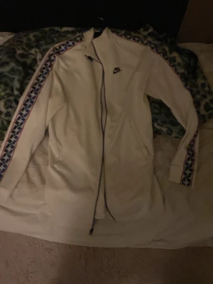 Vit Nike ziptröja med blå detaljer S - Säljer en vit Nike ziptröja i storlek S med blå och röda detaljer längs ärmarna och stort Nike-tryck på ryggen. Tröjan har hel dragkedja, hög krage och är tillverkad i ett mjukt syntetmaterial. Perfekt för dig som gillar sportig stil.