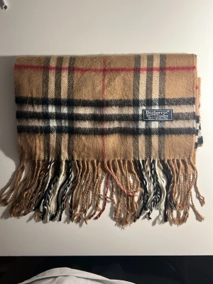 Rutig beige halsduk från Burberrys - Klassisk halsduk från Burberrys i 100% kashmir. Beige botten med rutigt mönster i svart, vitt och rött. Tidlös design med fransar i ändarna och mjuk, lyxig känsla. Perfekt accessoar för att lyfta din outfit.