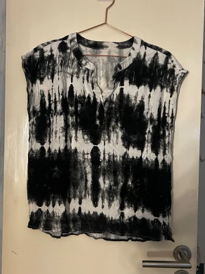 Svartvit tie-dye blus med v-ringning - Säljer en svartvit blus med tie-dye mönster och v-ringning. Blusen har korta ärmar och är tillverkad i ett tunt, mjukt material som känns skönt mot huden. Perfekt för dig som gillar en avslappnad och trendig stil.