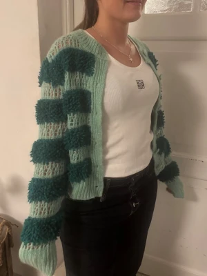 Kofta Zara - Unik stickad kofta i ljusgrönt med breda, mörkgröna fluffiga ränder. Koftan har rund halsringning, knappar framtill och ribbade muddar. Materialet ser ut att vara en mjuk ull- eller akrylblandning. Perfekt för dig som vill sticka ut med en cool och mysig look.