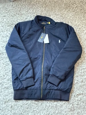 Marinblå bomberjacka från Polo Ralph Lauren - Snygg marinblå bomberjacka från Polo Ralph Lauren med klassisk passform. Jackan har ribbade muddar, dragkedja framtill och den ikoniska vita logon broderad på bröstet. Perfekt för dig som vill ha en stilren och tidlös look.