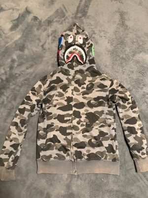Bape hoodie -   BAPE Shark Hoodie i grå camo. Full zip-hoodie med ikoniskt shark-motiv på huvan. Bra skick.   
