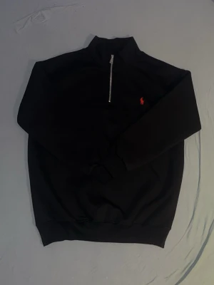 Svart half zip från Polo Ralph Lauren - Svart sweatshirt från Polo Ralph Lauren med klassisk röd logga broderad på bröstet. Tröjan har en halv dragkedja, hög krage och ribbade muddar. Perfekt för dig som gillar stilrena och tidlösa plagg.