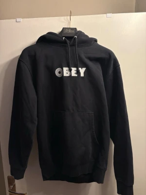Obey Font Logo Hoodie - Black - Size S - Selling this classic black hoodie from Obey Clothing. It features the iconic bold font logo embroidered/printed on the chest.  Condition: Excellent pre-owned condition, used sparingly. The fabric and logo are well-preserved.  Size: Small (S).  Details: Features a front kangaroo pocket and adjustable drawstring hood.  Authenticity: See photos for neck tags and branding.  A perfect streetwear staple that goes with everything!