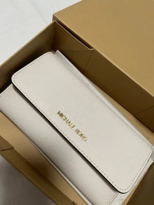 Vit plånbok från Michael Kors - Stilren vit äkta plånbok från Michael Kors i skinn med guldfärgad logga på framsidan. Plånboken har flera kortfack, ett fönster för ID och praktisk knappstängning, aldrig använd. Orginal box tillkommer❤️