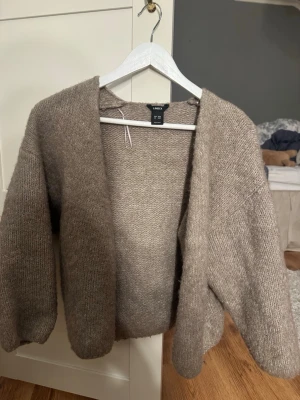 Beige stickad kofta från Lindex - Mysig beige stickad kofta från Lindex med öppen front och avslappnad passform. Koftan har långa ärmar och är gjord i ett mjukt, fluffigt material som känns skönt mot huden. Perfekt att slänga över en t-shirt eller topp för extra värme. Använd 2-3 gånger XS/S