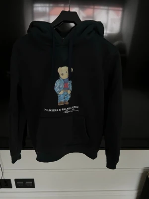 Marinblå hoodie Polo Bear Ralph Lauren - Marinblå hoodie från Ralph Lauren med den ikoniska Polo Bear-trycket framtill. Hoodien har huva med dragsko och en stor magficka. Materialet ser ut att vara mjukt och bekvämt, perfekt för en avslappnad stil.