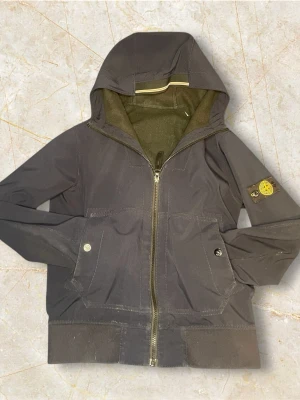 Mörkblå vindjacka från Stone Island - Säljer en mörkblå vindjacka från Stone Island med huva och dragkedja framtill. Jackan har två fickor med tryckknappar och den klassiska Stone Island-loggan på vänster ärm. Perfekt för dig som vill ha en clean sopig look. Har lite defekter på armarna
