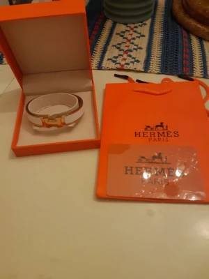 Vitt Hermès armband med guldspänne - Säljer ett stilrent vitt armband från Hermès med elegant guldspänne i klassisk H-form. Armbandet är gjort i läder och kommer i originalförpackning med tillhörande Hermès-påse. Perfekt accessoar för dig som gillar lyxiga detaljer.