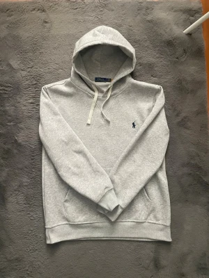 Grå hoodie från Polo Ralph Lauren - Snygg ljusgrå hoodie från Polo Ralph Lauren med klassisk huva och vita dragsnören. Tröjan har känguruficka framtill och den ikoniska mörkblå loggan broderad på bröstet. Tillverkad i mjuk bomullsblandning som känns skön mot huden.