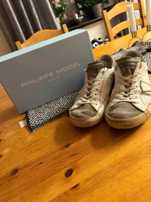 Philippe Model vita sneakers i skinn - Snygga vita sneakers från Philippe Model med grå detaljer och beige inslag. Skorna har klassisk snörning, rund tå och är tillverkade i skinn och mocka. Perfekta för dig som gillar stilrena och tidlösa sneakers med en exklusiv känsla. Bara att skriva om du har några frågor.