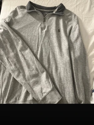 Grå halvzip från Polo Ralph Lauren - Snygg grå långärmad halvzip-tröja från Polo Ralph Lauren. Tröjan har en liten broderad logga på bröstet, hög krage med dragkedja och är tillverkad i mjukt bomullsmaterial. Perfekt för en avslappnad och stilren look. Nypris 2200