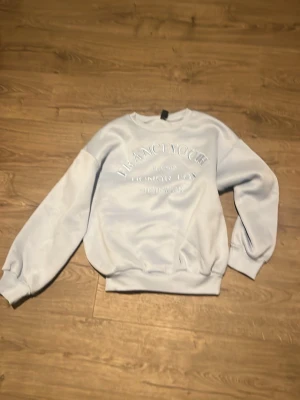 Ljusblå  sweatshirt från Shein - Ljusblå sweatshirt från Shein med broderad text framtill. Tröjan har rund hals, ribbade muddar vid ärmslut och nederkant samt en avslappnad passform. Perfekt för en chill och casual stil.