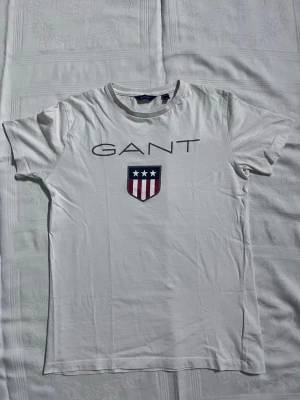 Vit GANT t-shirt med logga - Vit t-shirt från GANT med stor logga och sköldtryck framtill. Klassisk rund halsringning och korta ärmar. Tillverkad i mjuk bomull som känns skön mot huden. Perfekt för en clean och stilren look. 