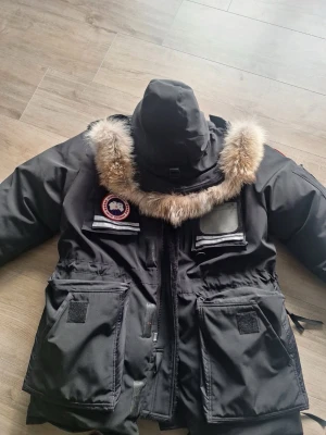 Svart dunjacka från Canada Goose - Säljer en svart dunjacka från Canada Goose med stor huva och äkta päls runt kragen. Jackan har flera praktiska fickor, tydlig logotyp på bröstet och robust dragkedja. Perfekt för kalla vinterdagar och har en klassisk, rymlig passform. PS. Jackan är stor i storleken