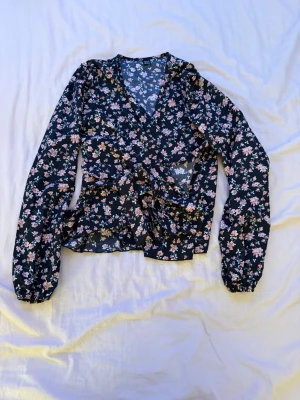 Blommig svart blus med knytband - Svart blus med blommigt mönster i rosa/vita toner. Lätt och luftig modell med långa ärmar och knytband framtill. Använd fåtal gånger, i mycket bra skick utan fläckar eller hål. Perfekt till både vardag och fest 