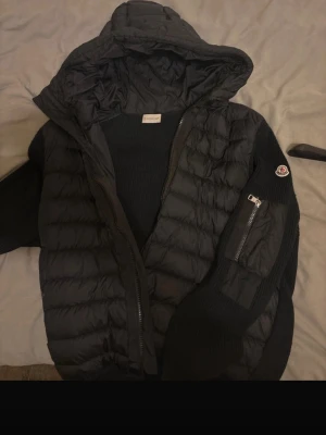 Svart Moncler pufferjacka med huva - Säljer en svart pufferjacka från Moncler med stickade ärmar och huva. Jackan har dragkedja framtill och en extra ficka med dragkedja på ärmen. Materialmix av ull och polyester ger både värme och stil. Perfekt för kyliga dagar.