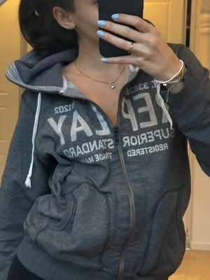 Mörkgrå zip-hoodie från Replay Superdry - Mörkgrå hoodie från Superdry med dragkedja framtill och vit text på bröstet. Tröjan har huva med vita dragsnören och två fickor framtill. Insidan av huvan är ljusgrå. Perfekt för en avslappnad och sportig stil.