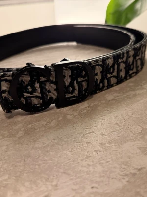 Märkes bälte - Säljer ett svart bälte  med ikoniskt monogrammönster och stilren metallspänne. Bältet är gjort i läder och textil, perfekt för att lyfta vilken outfit som helst med en lyxig touch.