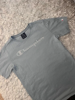 Ljusblå Champion t-shirt - Säljer en ljusblå t-shirt från Champion med klassisk logga i vitt framtill och liten logga på ärmen. T-shirten har rund hals och korta ärmar, perfekt för en avslappnad stil. Tillverkad i mjuk bomull för skön komfort.