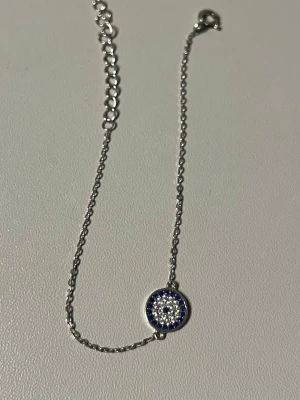 Silverarmband med blått cirkelhänge - Snyggt silverfärgat armband med ett runt hänge dekorerat med små vita och blå stenar i ett mönster som påminner om ett öga. Tunt kedjearmband med justerbar längd och karbinlås. Perfekt för dig som gillar stilrena men ändå unika detaljer. Aldrig använt 