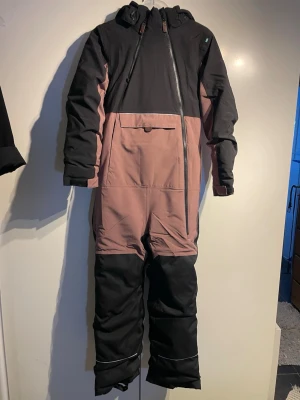 Overall, Svart och lila i stl 150 - En supervarm overall från Lindberg storlek 150. Har ett mjukt, stretchigt material. Den har ett värmande foder i fleece i kragen och en avtagbar huva som lossnar lätt om ditt barn skulle råka fastna.  - Vattentät. - Vattenpelare: 15000 mm. - Reflekterande detaljer. - Fothällor. - Två dragkedjor. - Ficka på magen. - Elastiska muddar. Nypris 1200:- 