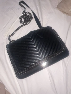 Svart quiltad axelväska med kedja - Snygg svart axelväska med quiltad design och silverfärgad kedjerem. Väskan har ett elegant mönster i v-format och detaljer i metall runt kanterna. Perfekt för dig som vill ha en stilren och trendig accessoar.