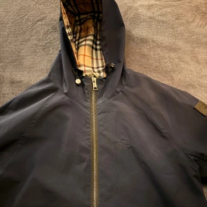 Marinblå vindjacka från Burberry - Snygg marinblå vindjacka från Burberry med klassiskt rutigt foder i huvan. Jackan har dragkedja framtill, två fickor med dragkedja och justerbar huva med snörning. Perfekt för blåsiga dagar och stilren look.