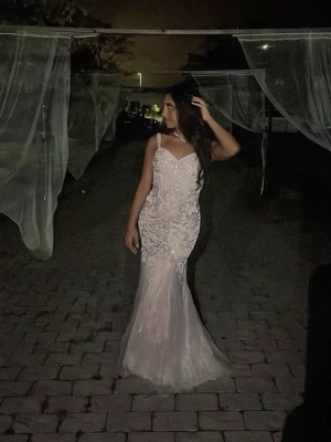 Vit långklänning med spetsdetaljer - Elegant vit långklänning med smala axelband och v-ringning. Klänningen har vackra spetsdetaljer och figurnära passform som övergår i en lätt utsvängd kjol. Perfekt för speciella tillfällen.