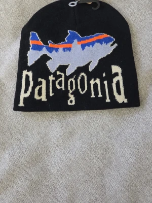 Svart mössa från Patagonia med fiskmotiv - Svart mössa från Patagonia med stor fisk i blått, vitt och orange samt logotypen i vitt framtill. Stickad i mjukt material och har en avslappnad passform. Perfekt för dig som gillar outdoor och streetwear.