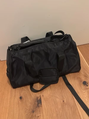 Svart sportväska från MM Sports - Säljer en svart sportväska från MM Sports med både handtag och justerbar axelrem. Väskan är tillverkad i slitstark polyester och har dragkedja upptill. Perfekt för gymmet eller träningstillfällen.