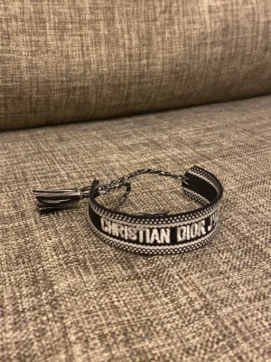 Christian Dior svart tygarmband - Snyggt svart och vitt tygarmband från Christian Dior med texten 'CHRISTIAN DIOR J'ADIOR' broderat runt om. Armbandet har justerbar knytning med tofsar i ändarna och en cool, trendig look som passar perfekt till din outfit.