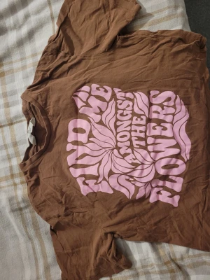 Brun t-shirt med rosa tryck - Cool brun t-shirt med stort rosa retrotryck på framsidan där det står 'Find me amongst the flowers' i vågig text. T-shirten har klassisk passform och korta ärmar, perfekt för en avslappnad stil.