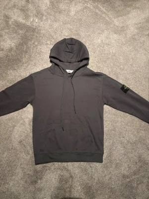 Grå hoodie från Stone Island - Snygg grå hoodie från Stone Island med klassisk logotyp-patch på vänster ärm. Hoodien har huva med dragsko och en stor magficka framtill. Tillverkad i mjuk bomull för en skön och avslappnad stil.