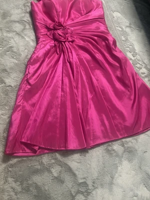 Rosa axelbandslös kort klänning - Axelbandslös kort klänning i glansigt rosa tyg med draperad midja och dekorativ blomdetalj framtill. Kjolen har ett fint fall och materialet ger en lyxig känsla. Perfekt för dig som vill sticka ut med en färgstark look.