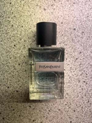 Yves Saint Laurent parfymflaska - Stilren parfym från Yves Saint Laurent i en transparent, fyrkantig glasflaska med svart, räfflad kork. Flaskan har en metallisk etikett med loggan på framsidan och känns både modern och lyxig. Perfekt för dig som gillar exklusiva dofter och snygg design på badrumshyllan. 60ml