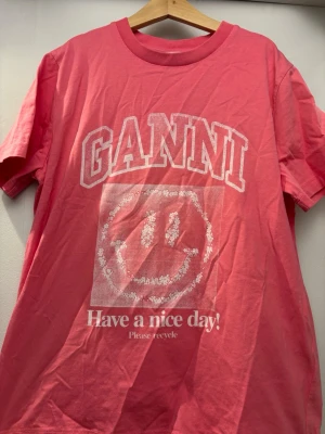 Rosa GANNI t-shirt med smileytryck - Cool rosa t-shirt från GANNI med stort vitt tryck framtill, där det står 'GANNI' och har en smiley samt texten 'Have a nice day! Please recycle'. Klassisk rund hals och korta ärmar. Perfekt statement-plagg för dig som gillar färg och grafiska prints. Plagget är bara använt ett fåtal gånger och är i ett mycket bra skick
