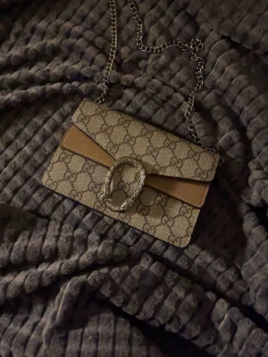  beige axelväska med kedja - Säljer en beige axelväska  med klassiskt GG-mönster, silverfärgad kedjerem och snygg detalj i form av ett dekorativt spänne framtill. Väskan har bruna skinnpartier och är perfekt för dig som gillar lyxiga accessoarer. Pris kan diskuteras 