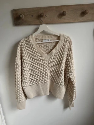 Beige stickad tröja från Kappahl  - Säljer en beige stickad tröja från Kappahl med djup v-ringning och grovt strukturmönster. Tröjan har breda ribbade muddar vid ärmslut och nederkant samt en avslappnad passform. Perfekt för lager på lager och riktigt mysig.